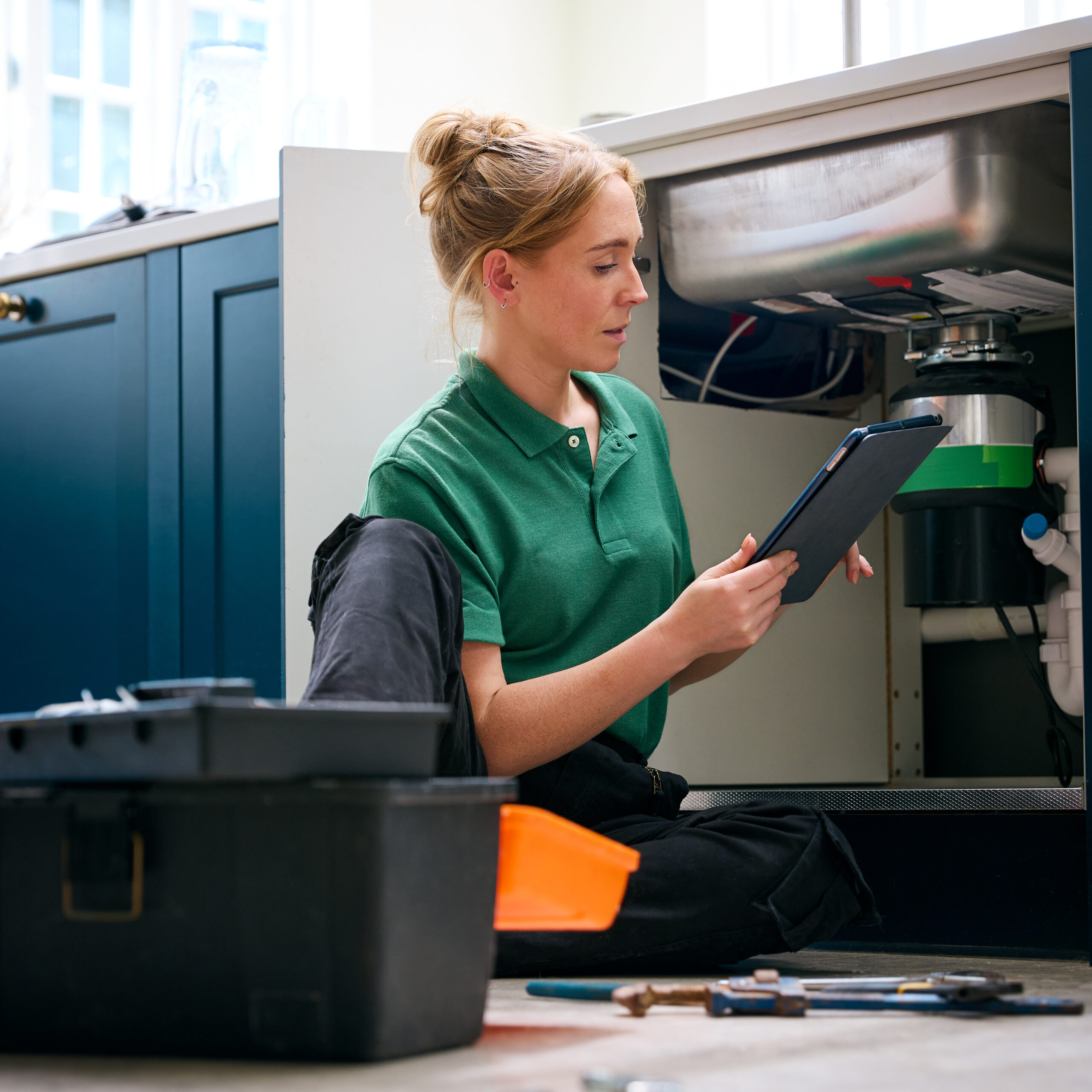 Female Plumber With Digital Tablet Fixing Waste Disposal Unit In Domestic Kitchen Sink